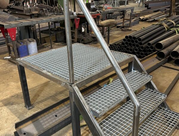 Tērauda pakāpienu konstrukcija ar metinātu rāmi un roku balstu industriālā darbnīcas vidē.